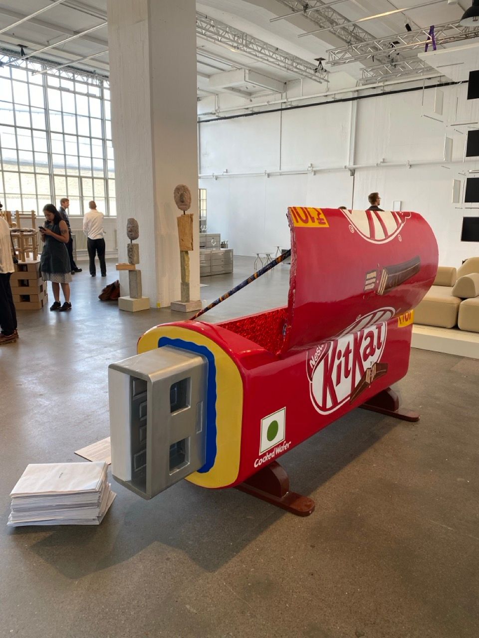 A large red Kit Kat bar sculpture with a USB plug on one end sits on the floor of an exhibition space.