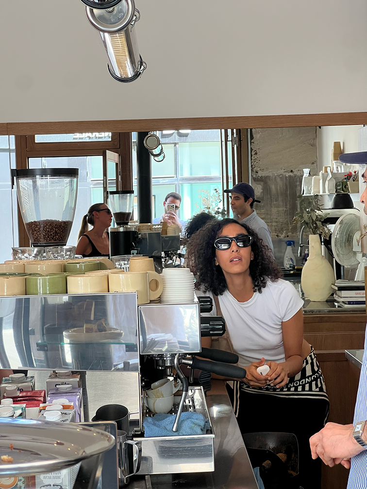 A woman with curly hair and sunglasses, wearing a white t-shirt, stands behind a coffee bar with an espresso machine, looking up.