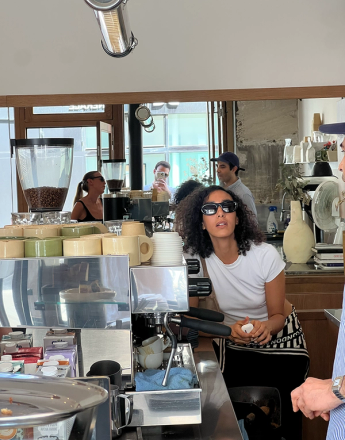 A woman with curly hair and sunglasses, wearing a white t-shirt, stands behind a coffee bar with an espresso machine, looking up.