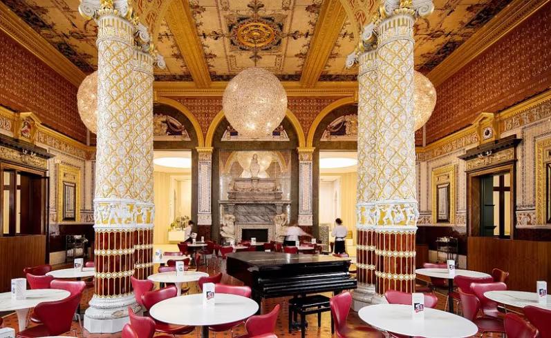 A grand, ornate cafe interior with decorative white and gold columns, a patterned ceiling, red chairs, spherical chandeliers, and a grand piano.