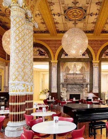 A grand, ornate cafe interior with decorative white and gold columns, a patterned ceiling, red chairs, spherical chandeliers, and a grand piano.