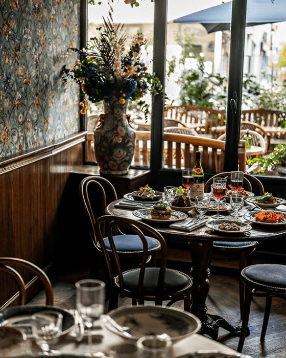 Restaurant interior with a dining table set with food and wine, a large floral centerpiece, and windows looking out to an outdoor patio.