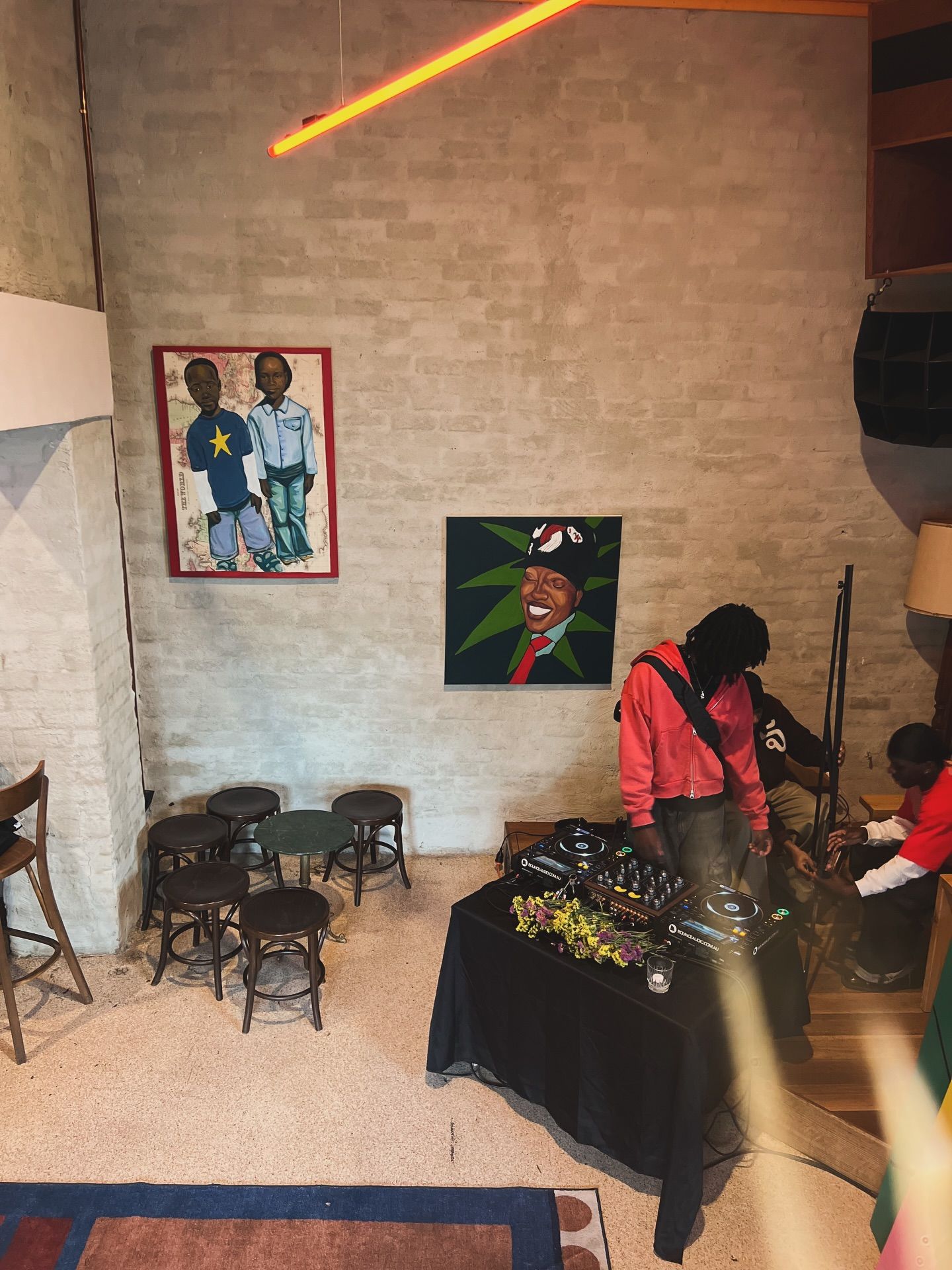 An overhead view of a room with a DJ setup, people working around it, and two paintings on a brick wall, illuminated by a neon light.