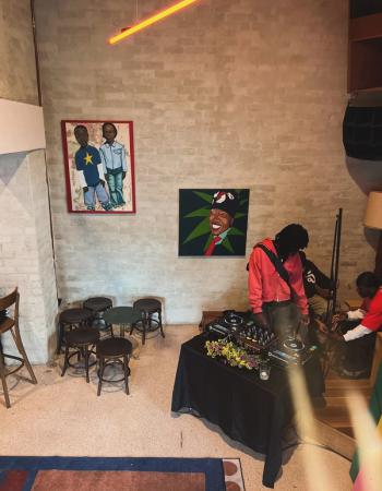 An overhead view of a room with a DJ setup, people working around it, and two paintings on a brick wall, illuminated by a neon light.