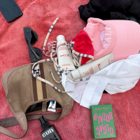 Beach day items scattered on a pink towel, including a brown bag, sunglasses, phone, sunscreen, a red bikini, a pink cap, and a green box.