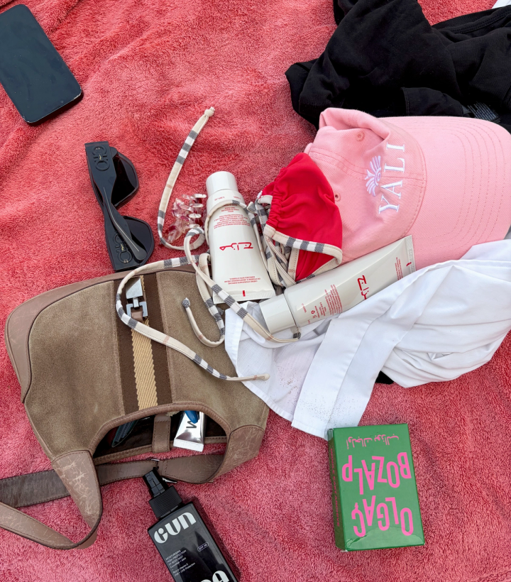 Beach day items scattered on a pink towel, including a brown bag, sunglasses, phone, sunscreen, a red bikini, a pink cap, and a green box.