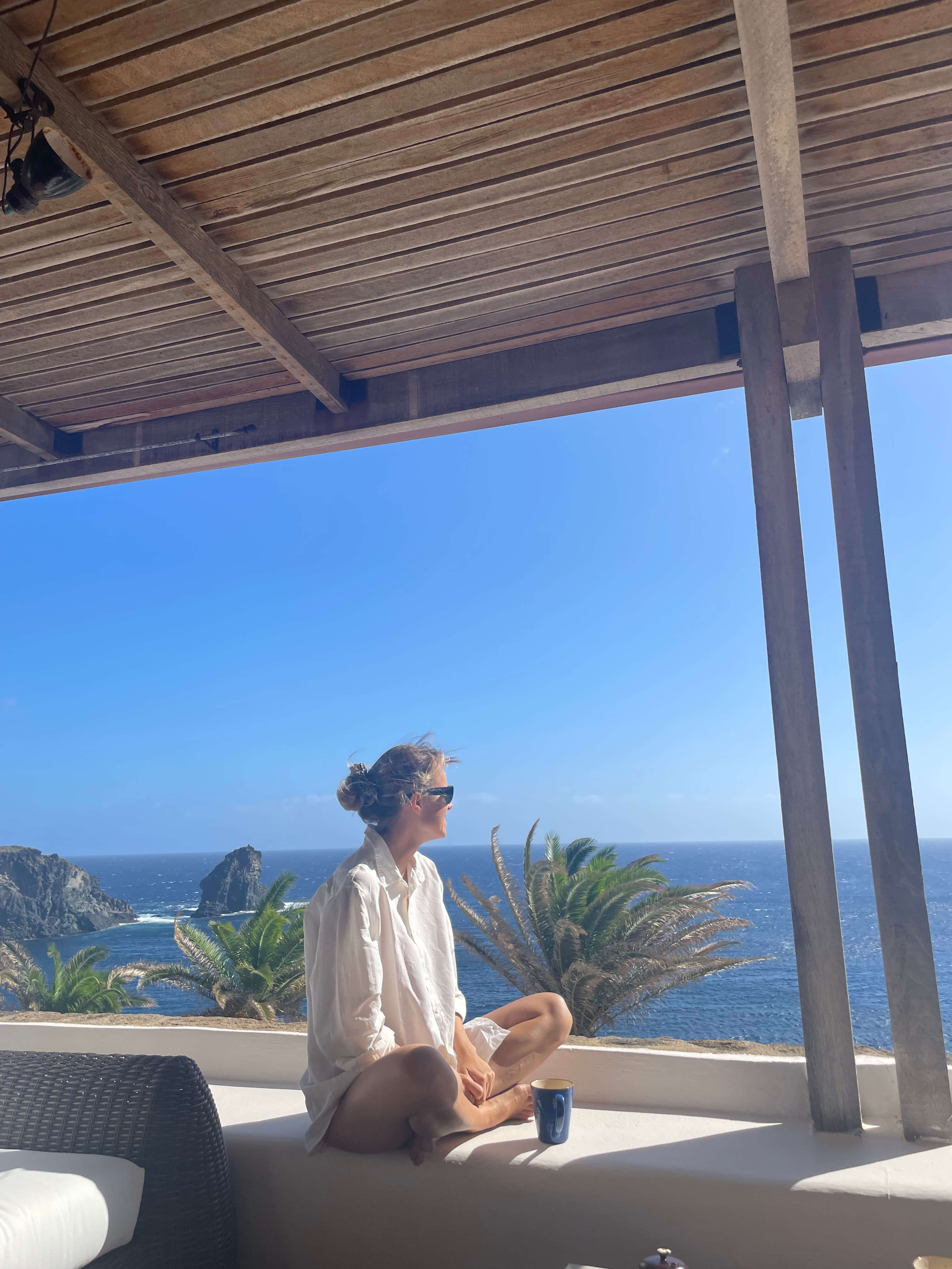 A woman sits on a sunny balcony ledge, looking out at a vast blue ocean with rocky cliffs and palm trees.