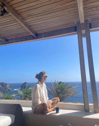 A woman sits on a sunny balcony ledge, looking out at a vast blue ocean with rocky cliffs and palm trees.