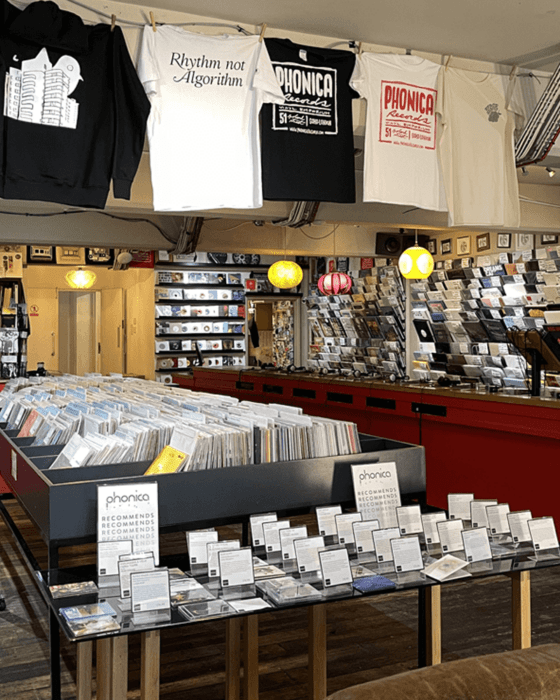 A vibrant record store interior with "Phonica Records" t-shirts hanging, including one reading "Rhythm not Algorithm," large bins filled with vinyl records, and display tables showcasing recommended albums.