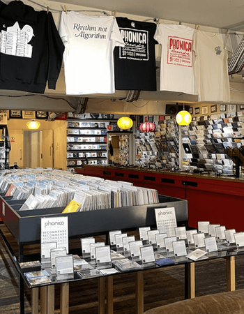 A vibrant record store interior with "Phonica Records" t-shirts hanging, including one reading "Rhythm not Algorithm," large bins filled with vinyl records, and display tables showcasing recommended albums.