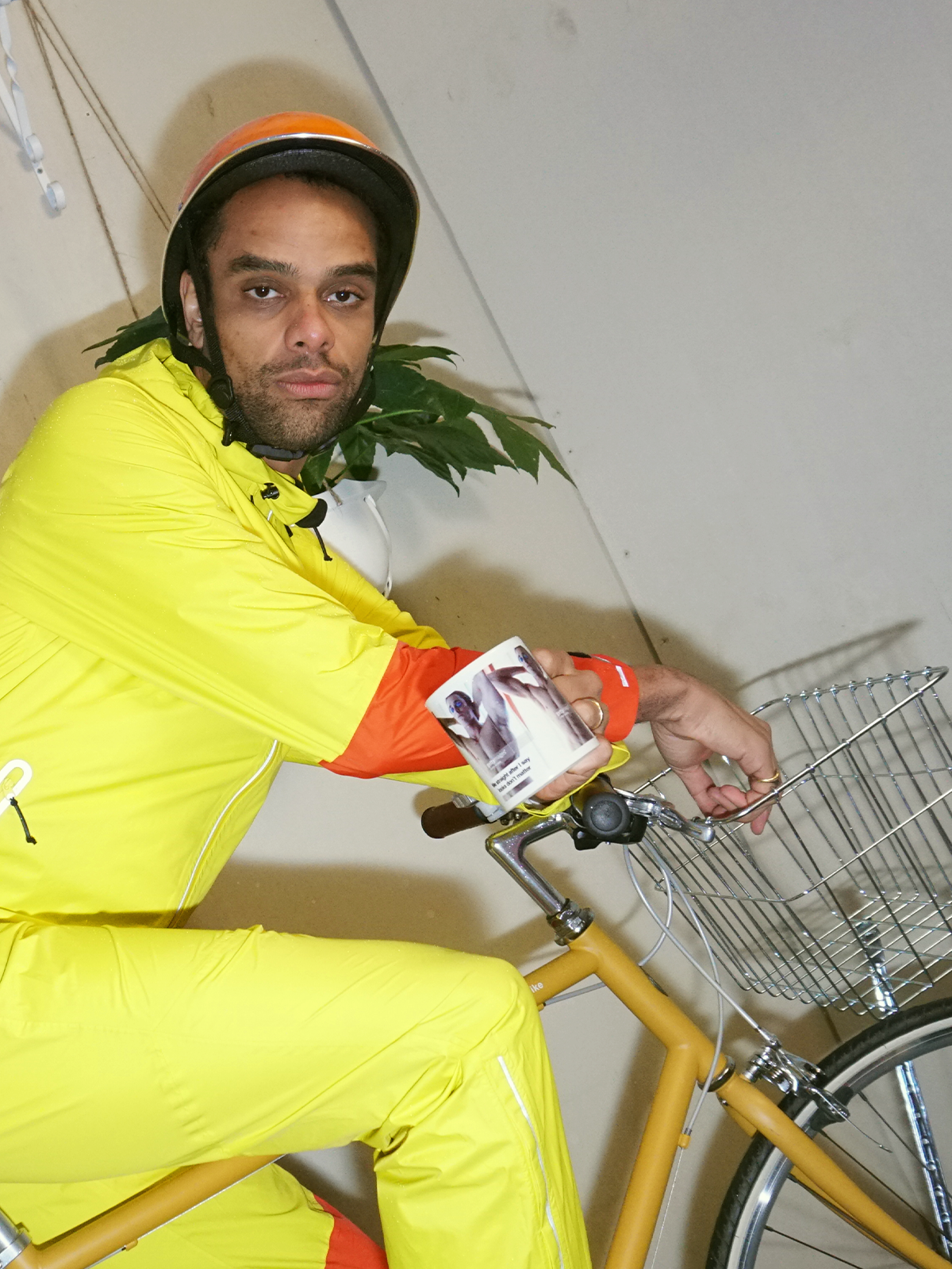 A man in a yellow jumpsuit and helmet sits on a yellow bicycle with a front basket, holding a mug.