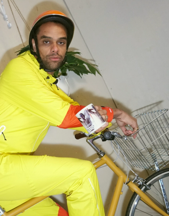A man in a yellow jumpsuit and helmet sits on a yellow bicycle with a front basket, holding a mug.