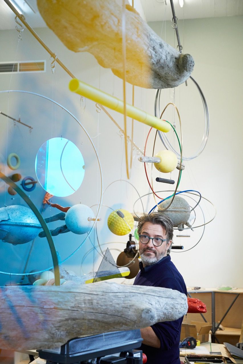 A bearded man with glasses stands in front of a complex, multi-component hanging sculpture.