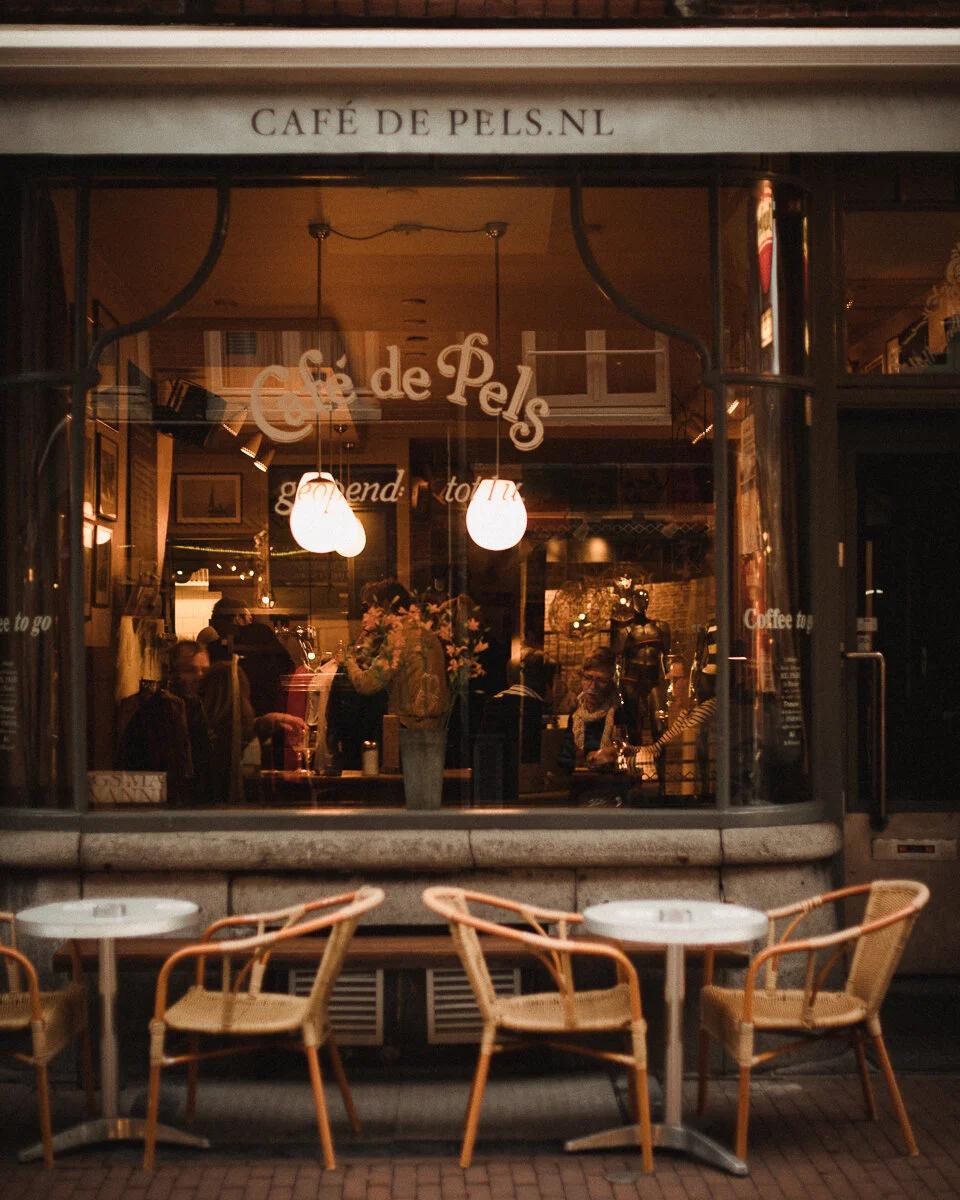 Exterior view of Café de Pels with outdoor tables and chairs, showing a warmly lit interior with people.