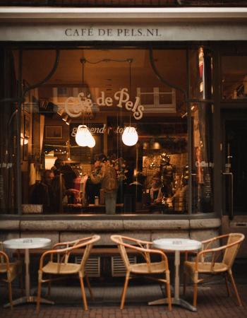 Exterior view of Café de Pels with outdoor tables and chairs, showing a warmly lit interior with people.