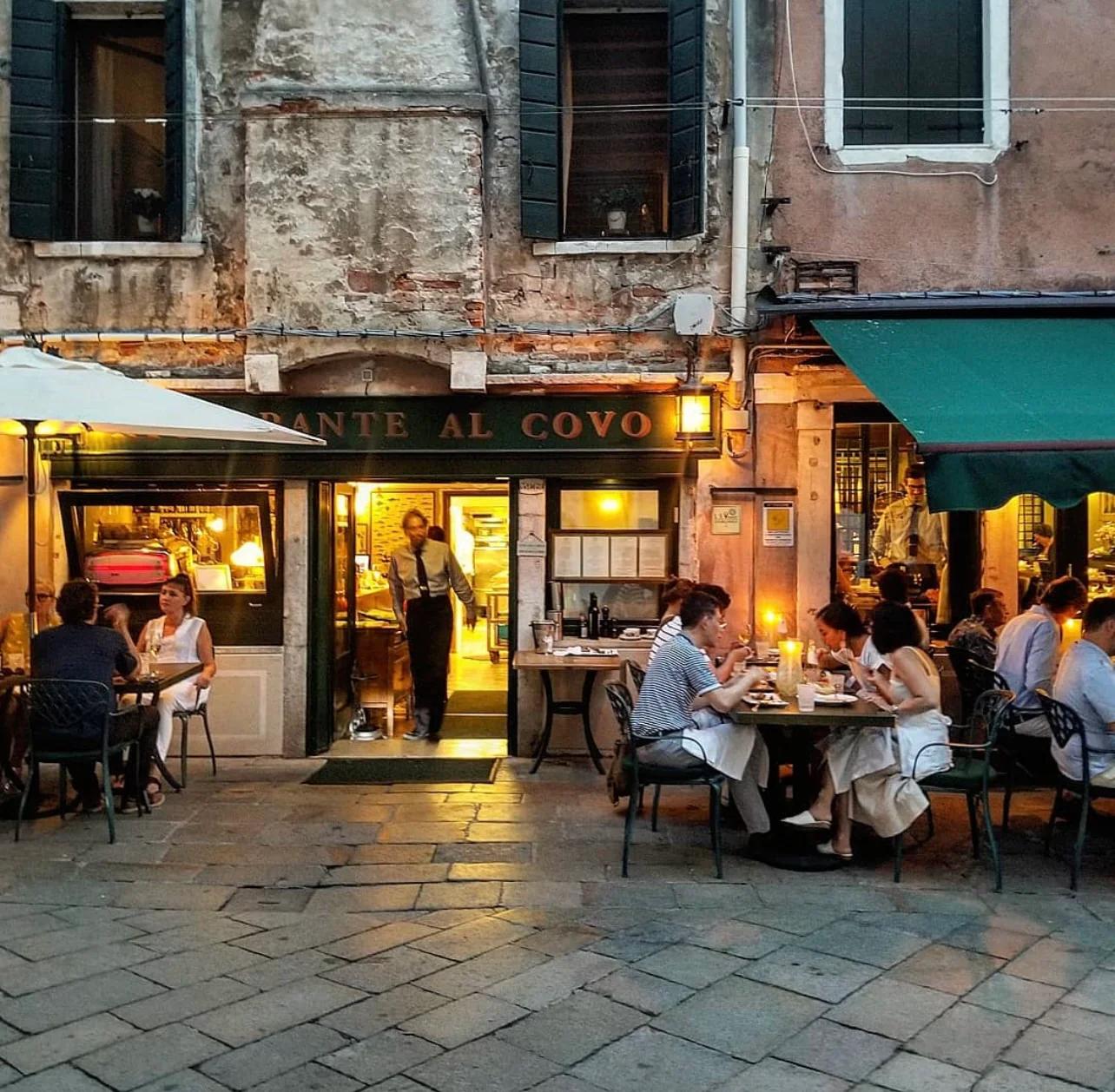Evening scene of people dining outdoors at the 'AL COVO' restaurant on a cobbled street, lit by warm lights.