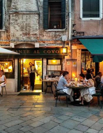 Evening scene of people dining outdoors at the 'AL COVO' restaurant on a cobbled street, lit by warm lights.