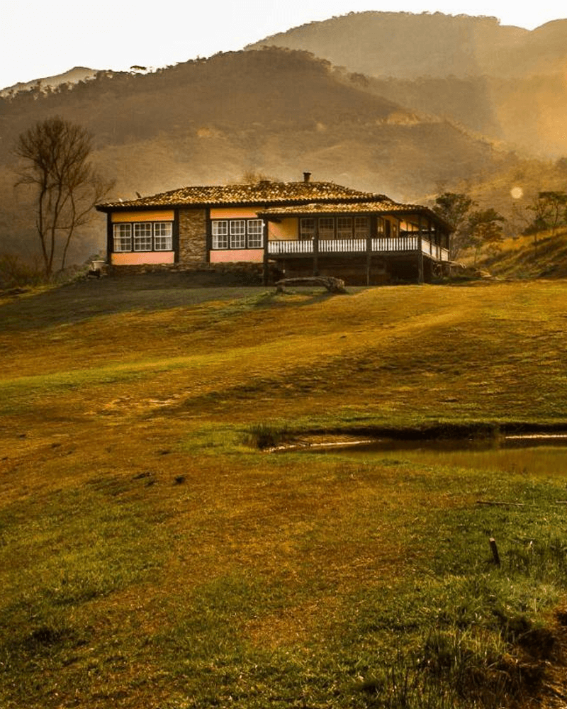A single-story house with a wide porch sits on a golden-hued grassy slope, with mountains in the background and a small pond in the foreground.