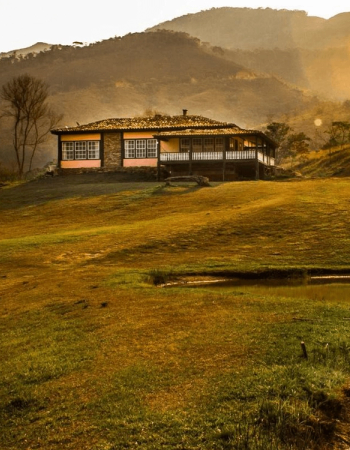 A single-story house with a wide porch sits on a golden-hued grassy slope, with mountains in the background and a small pond in the foreground.