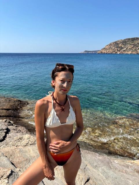A woman with a face mask on stands in a bikini on a rocky shore by clear blue sea under a sunny sky.
