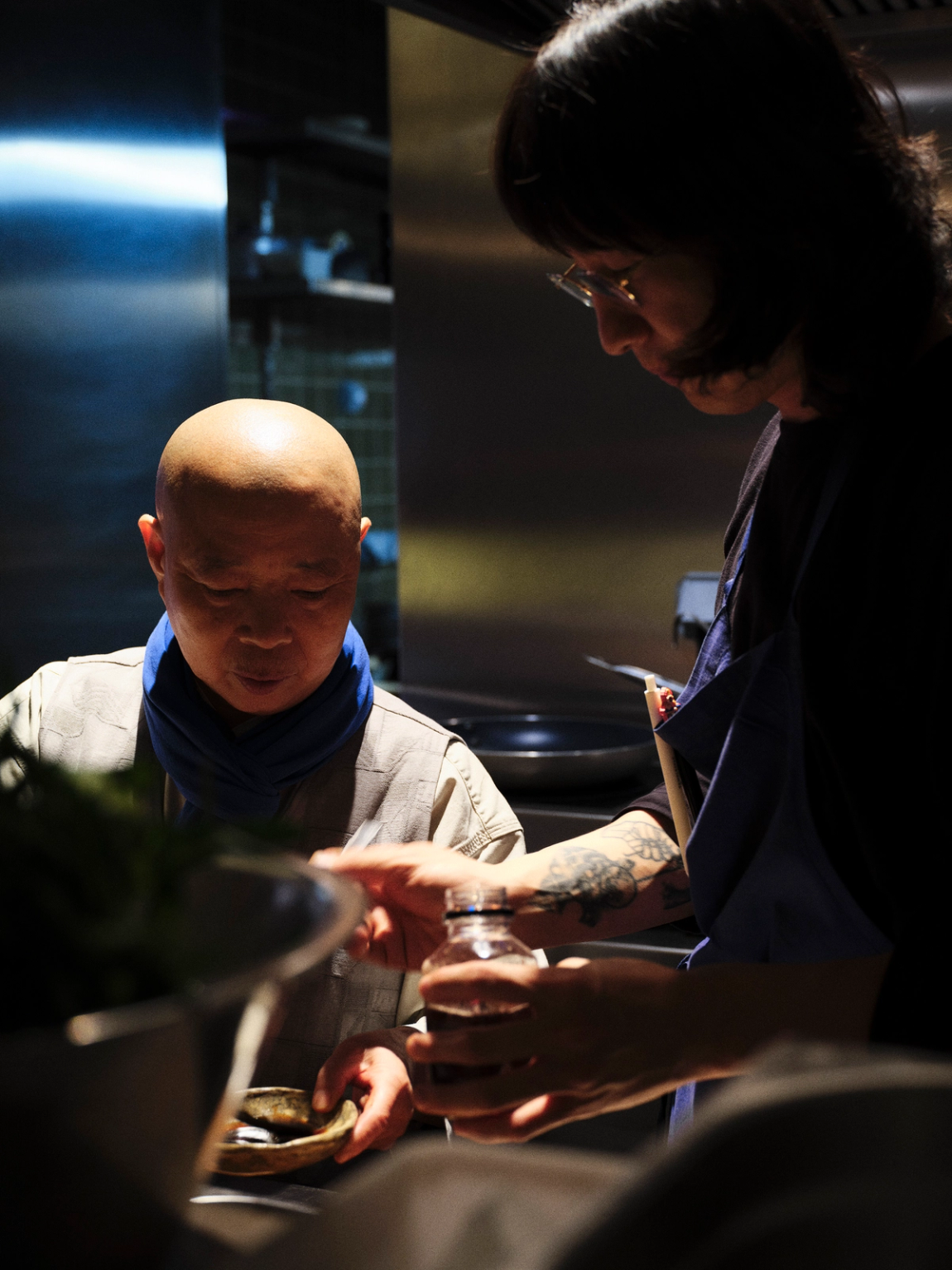 A bald chef in a blue scarf looks into a small bowl, while a tattooed person in an apron holds a bottle and gestures towards it.
