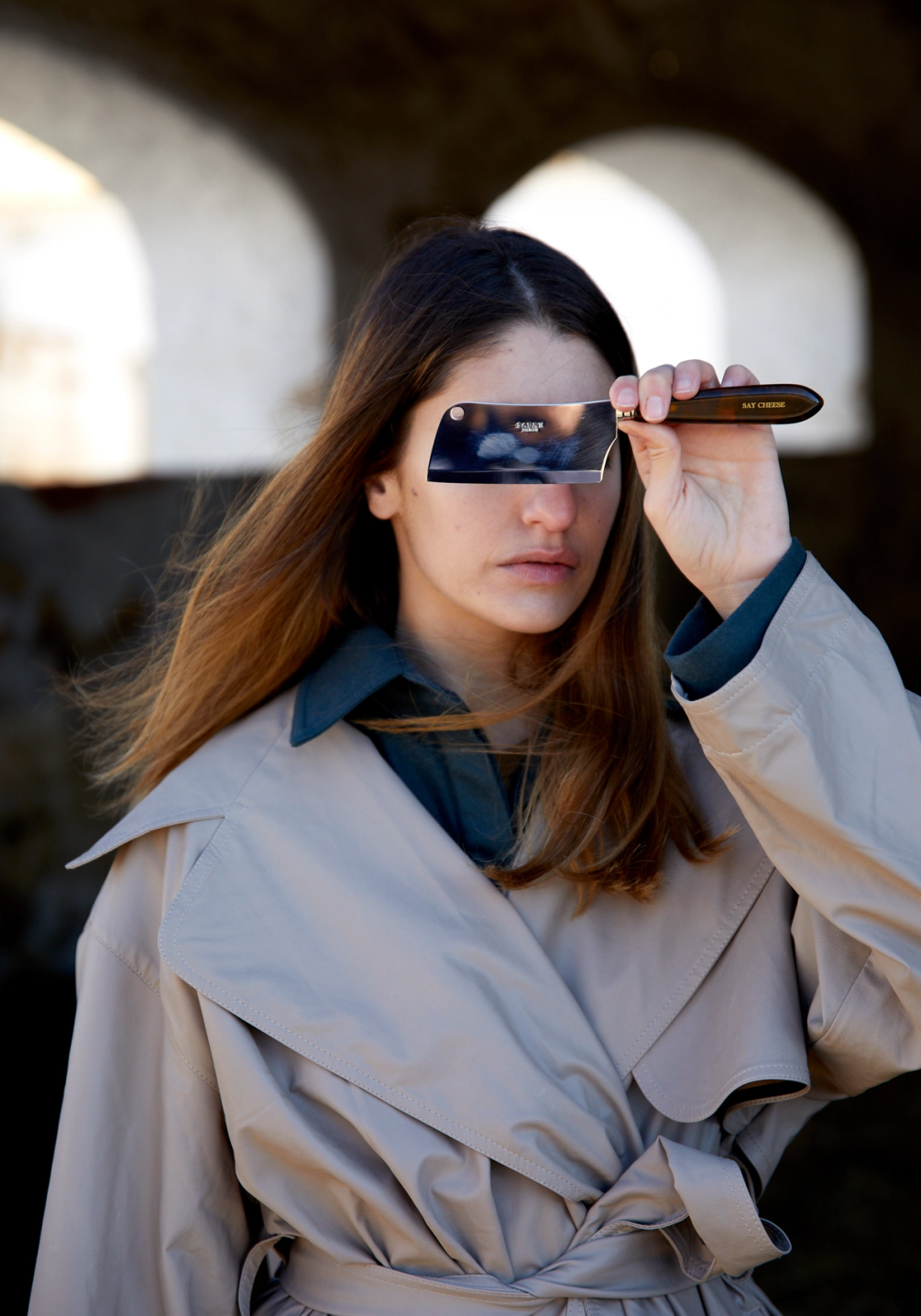 Woman in a trench coat holding a small cleaver over her right eye.