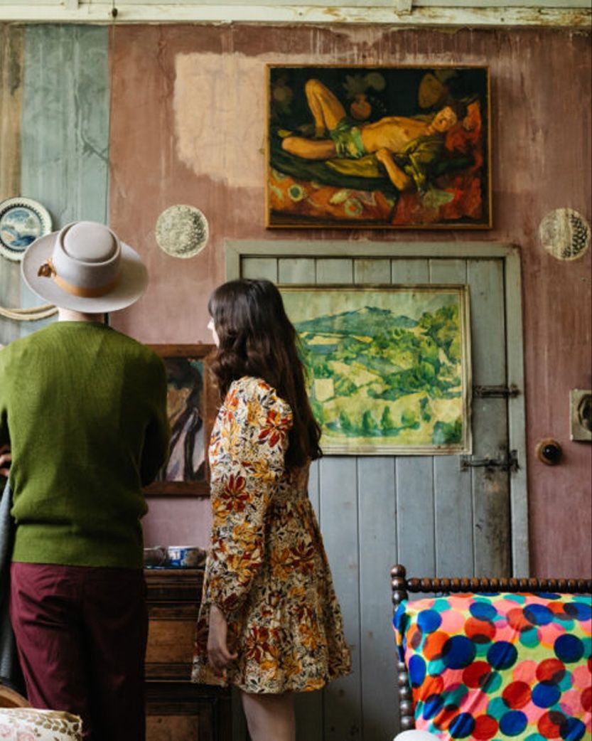 Two people from behind in a rustic room with pink walls, looking at a painting of a reclining figure.