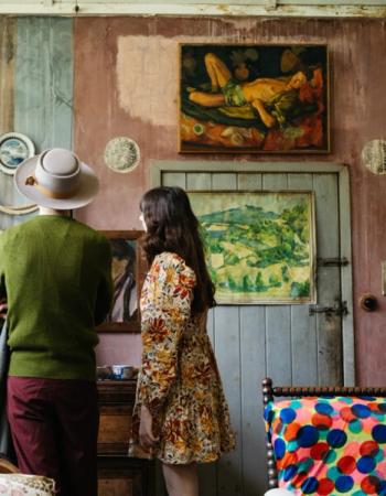 Two people from behind in a rustic room with pink walls, looking at a painting of a reclining figure.
