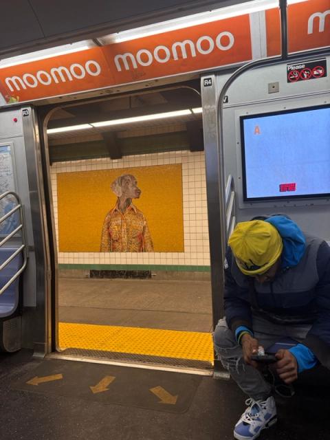 Inside a subway car with an overhead "moomoo" banner, a man in a yellow beanie crouches looking at his phone. The open door reveals a subway platform and a large mosaic of a dog in a shirt on a yellow wall.