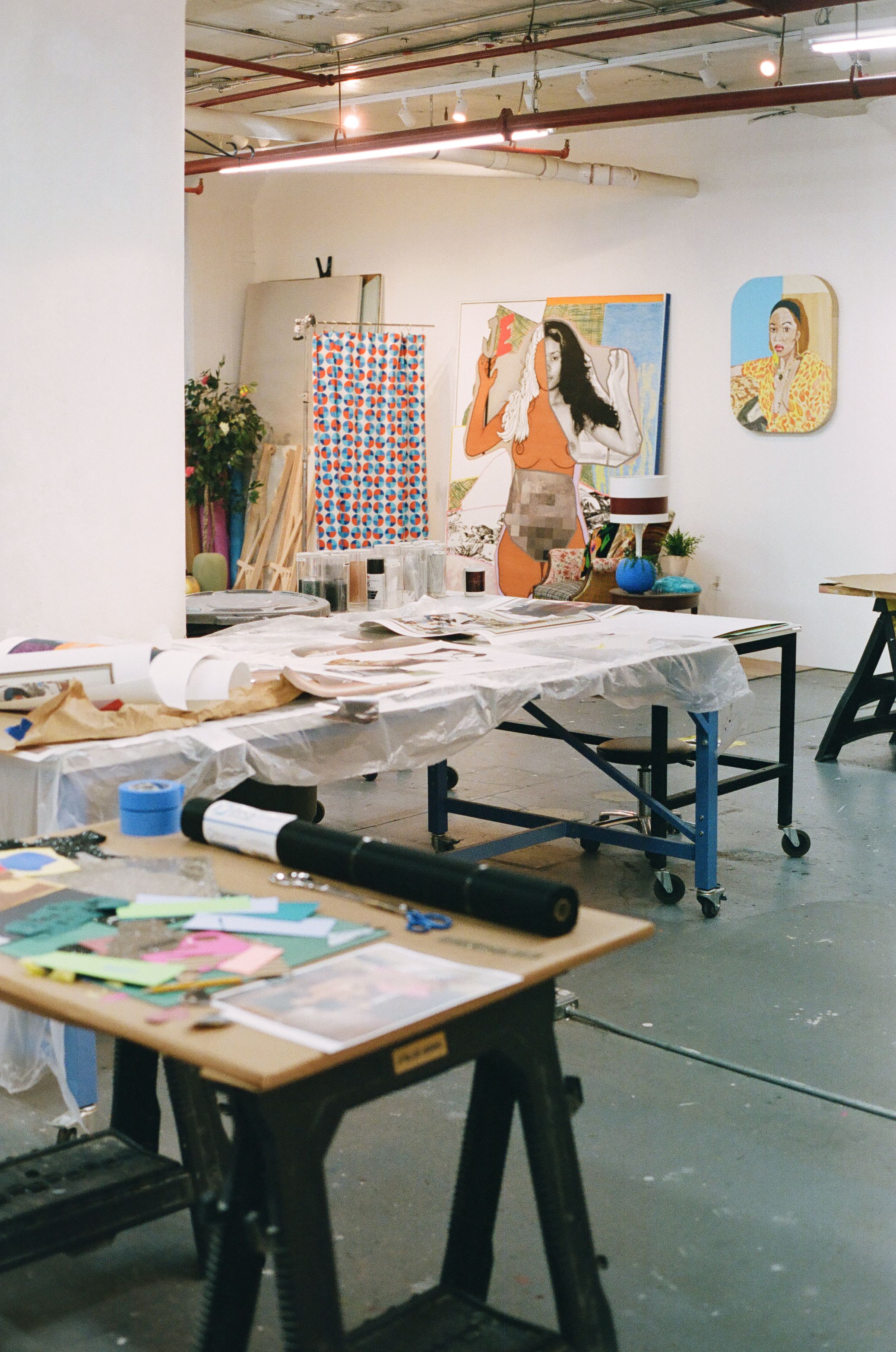 An art studio with work tables covered in supplies, and large paintings, including a pixelated female figure and a portrait, hanging on white walls.