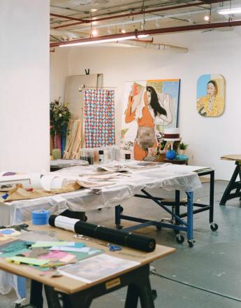 An art studio with work tables covered in supplies, and large paintings, including a pixelated female figure and a portrait, hanging on white walls.