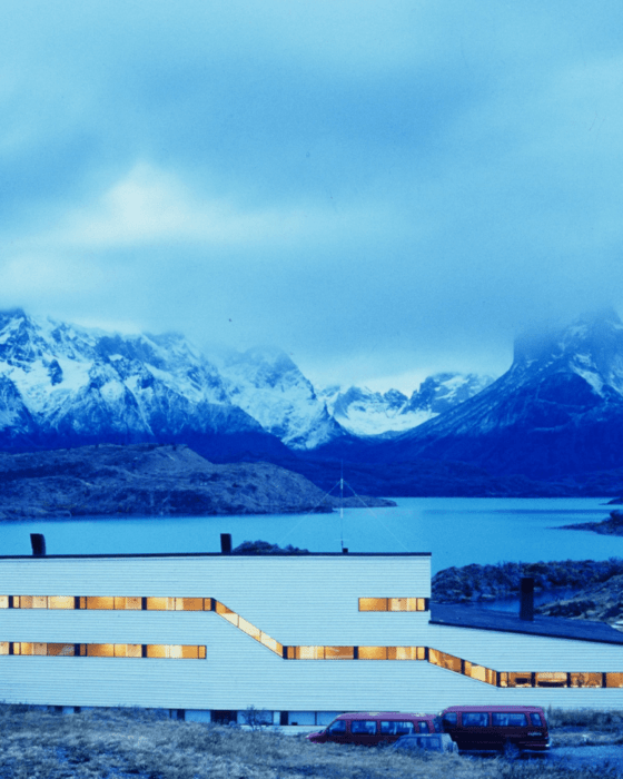 A modern white building with glowing windows and red vans, set against a blue lake and snow-capped mountains.