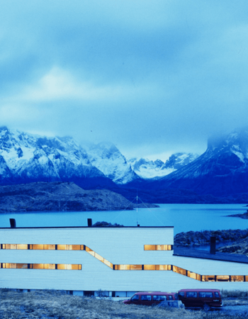 A modern white building with glowing windows and red vans, set against a blue lake and snow-capped mountains.