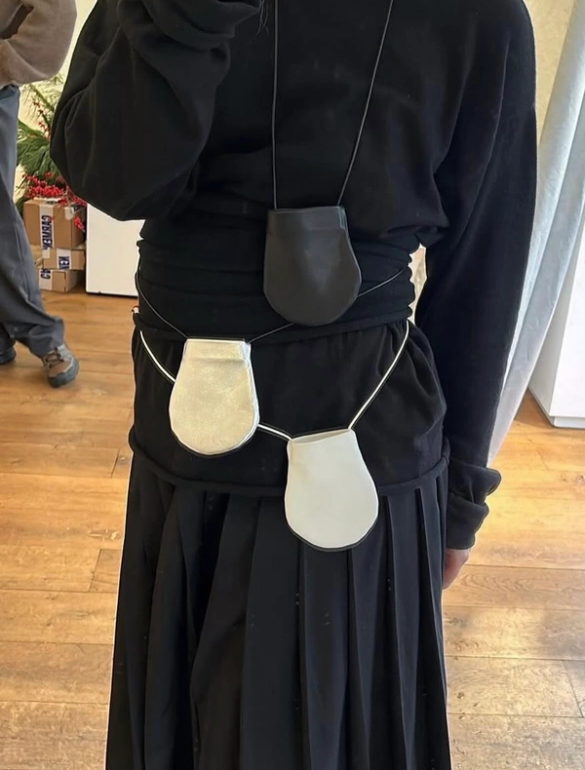 A person from behind wears a black top and pleated skirt, accessorized with a dark gray pouch around the neck and two light gray pouches connected around the waist.