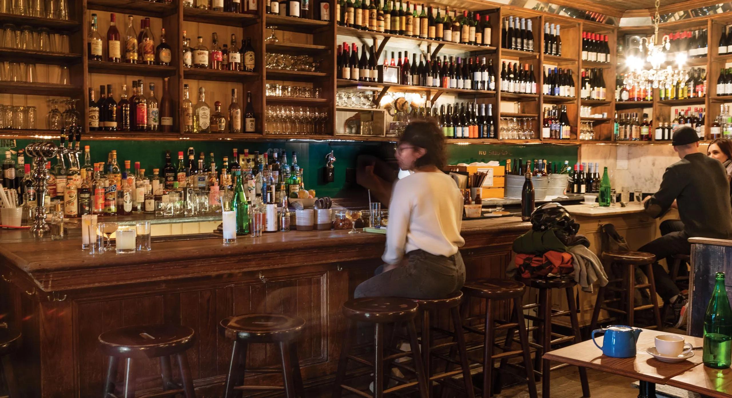 Warmly lit bar interior with a long wooden counter, shelves filled with liquor and wine bottles, and patrons seated at the bar.
