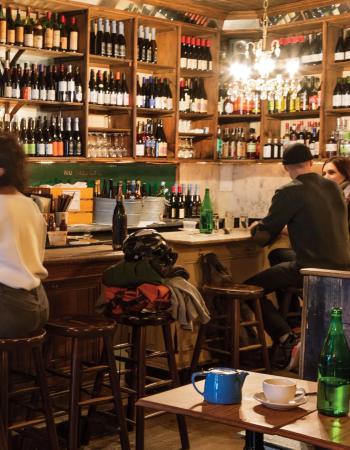 Warmly lit bar interior with a long wooden counter, shelves filled with liquor and wine bottles, and patrons seated at the bar.