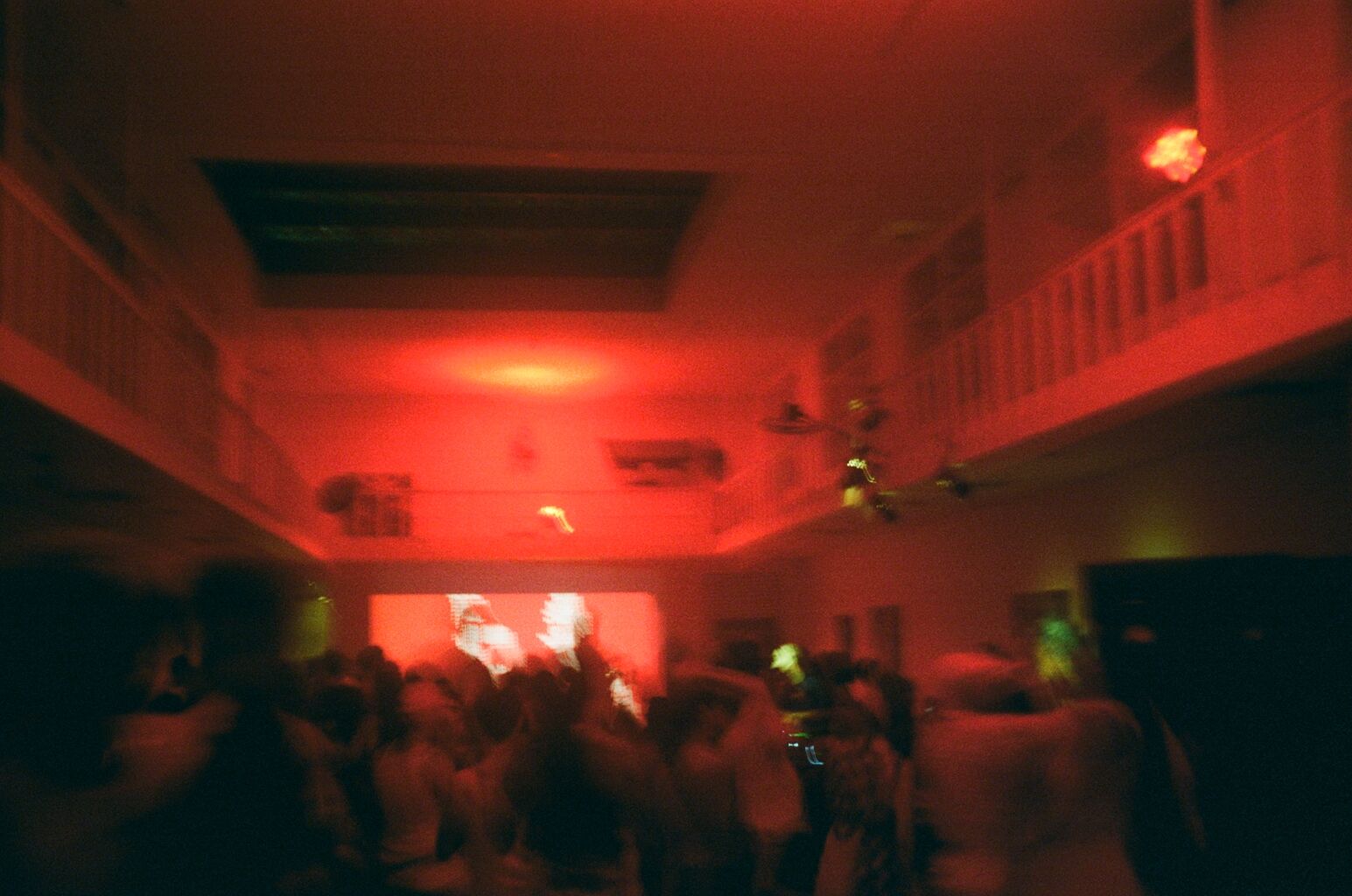 A blurry, red-lit image of a crowded room with a balcony and a projection screen.
