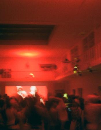 A blurry, red-lit image of a crowded room with a balcony and a projection screen.