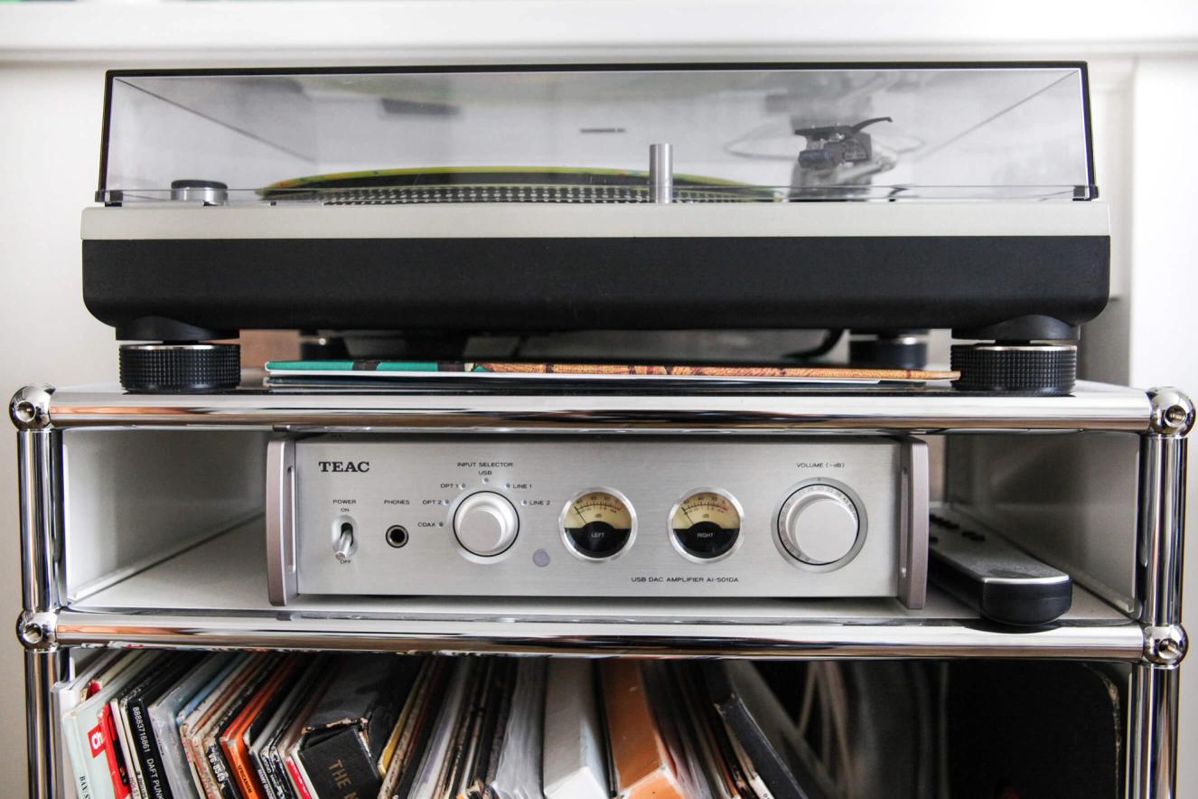 A turntable sits atop a TEAC audio amplifier, both on a modern metal shelving unit with vinyl records stacked on the shelf below.