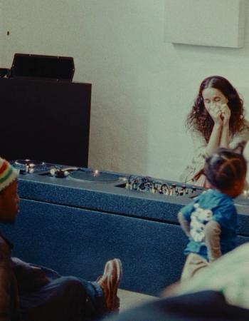 A man, child, and woman in a room with a DJ setup, the woman covering her mouth.