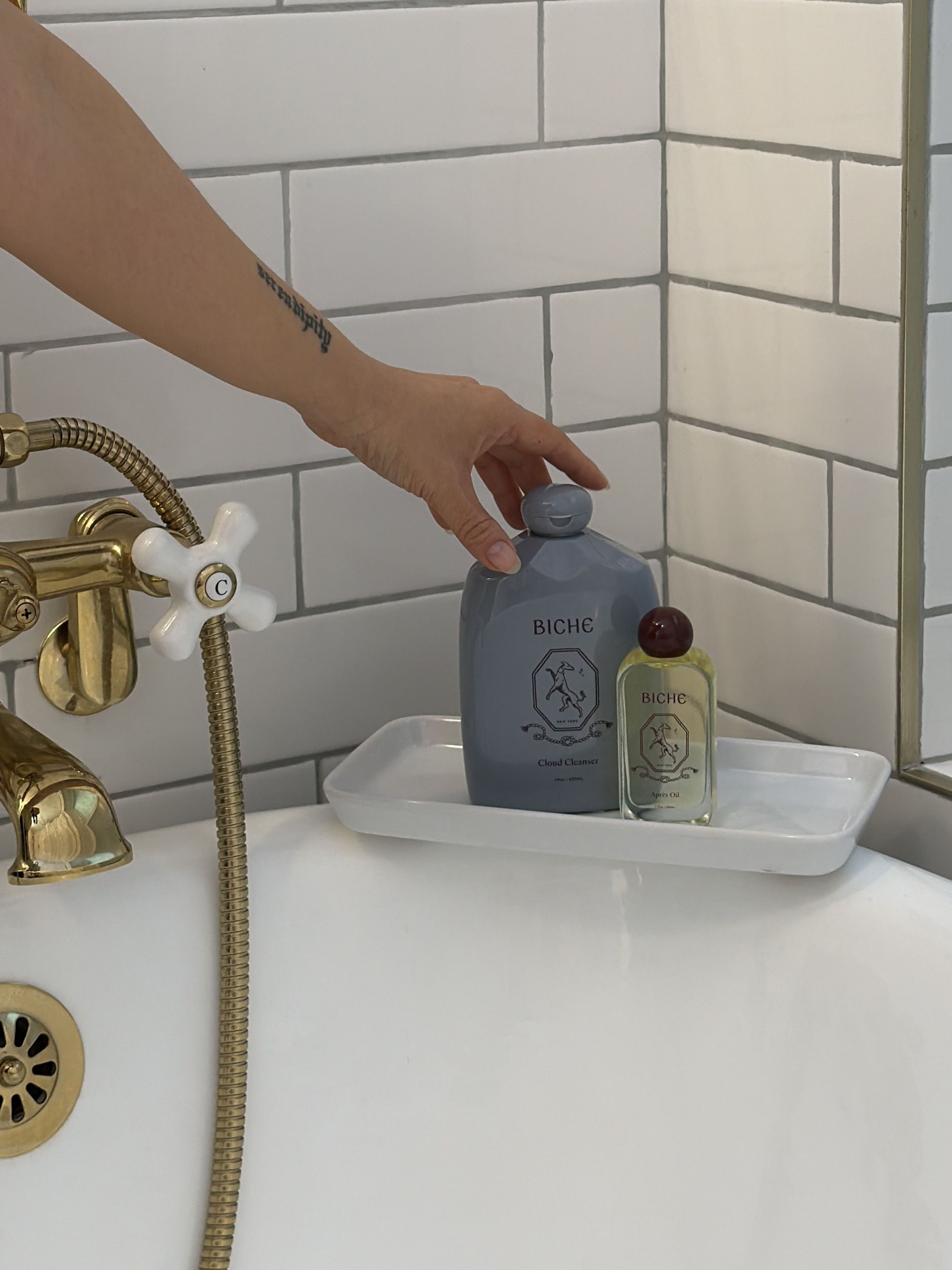 A hand reaches for a grey bottle on a white tray in a bathtub with brass fixtures.