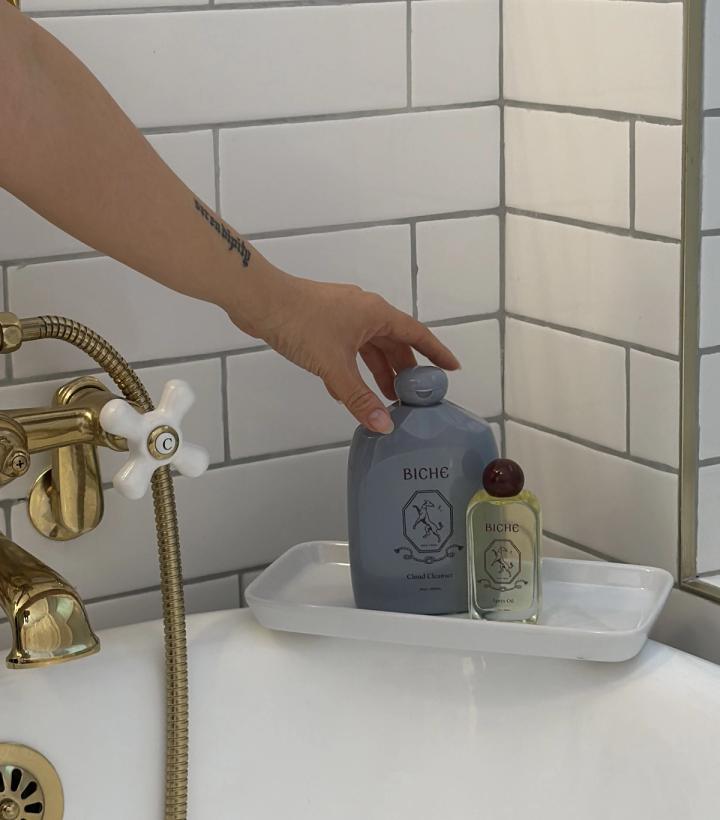 A hand reaches for a grey bottle on a white tray in a bathtub with brass fixtures.