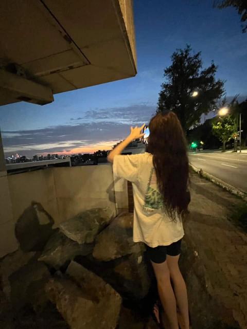 A person with long dark hair photographs a colorful sunset over a distant city from under a concrete overpass.