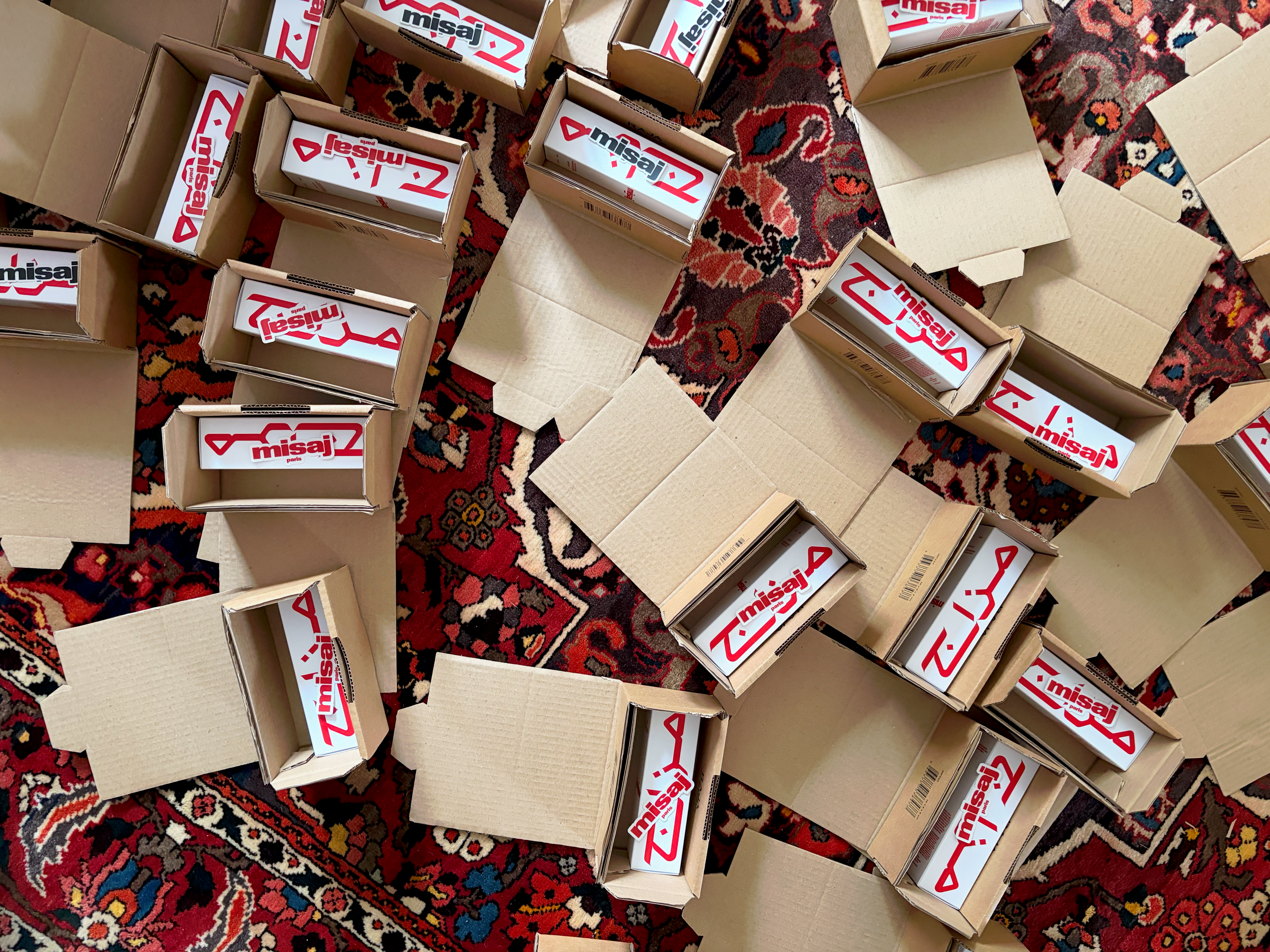 Numerous open cardboard boxes containing white products with red "misaj" text in English and Arabic are scattered on a patterned rug.