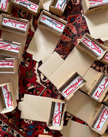 Numerous open cardboard boxes containing white products with red "misaj" text in English and Arabic are scattered on a patterned rug.