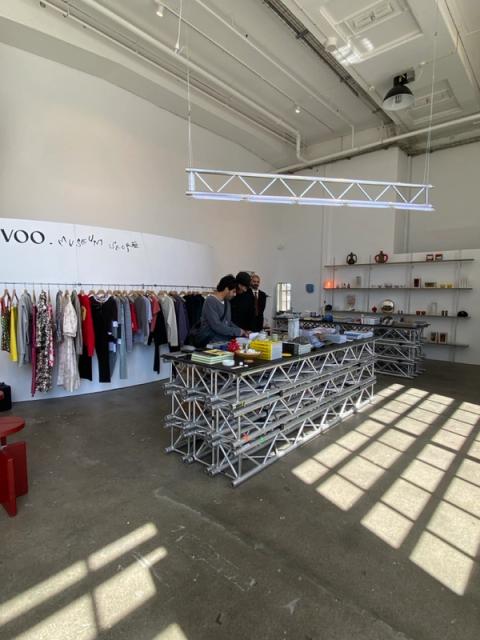 A modern shop, "VOO. Museum Shop," with clothes on a rack, two large metal truss tables displaying merchandise, and shelves of goods. Natural light casts shadows on the concrete floor.