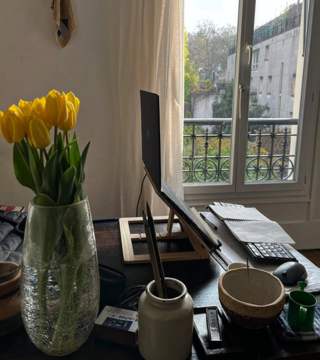 A desk with a laptop on a stand, a vase of yellow tulips, and other items, by a window showing trees and buildings.