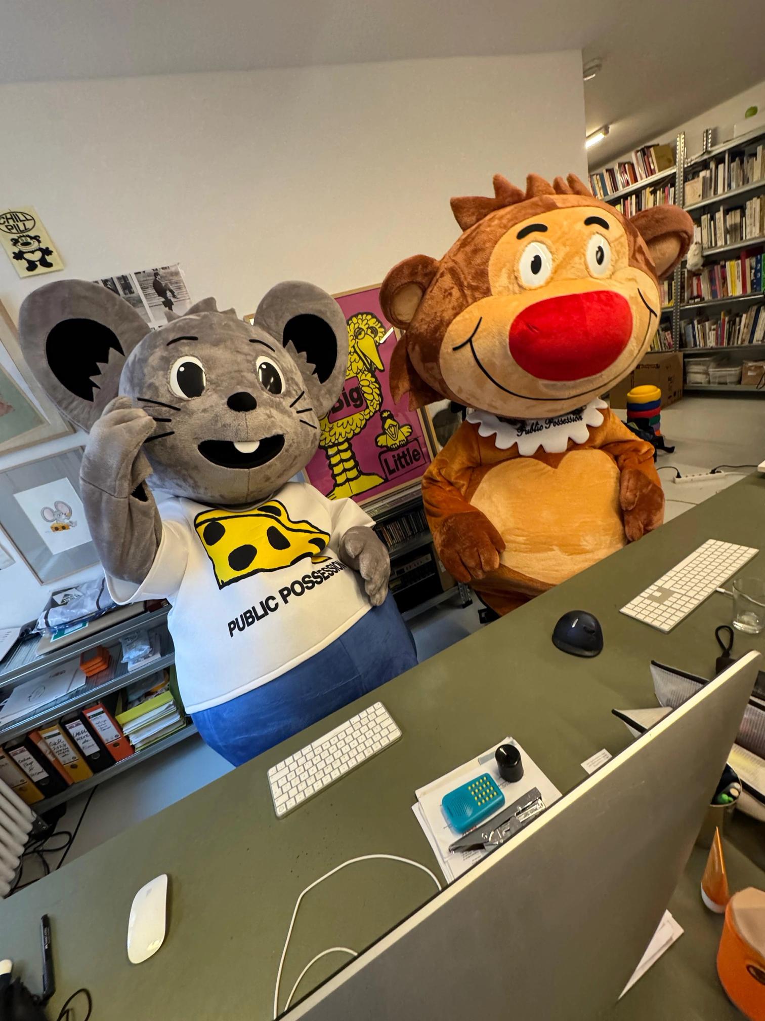 Two mascots, a gray mouse and a brown monkey, stand behind an office desk.
