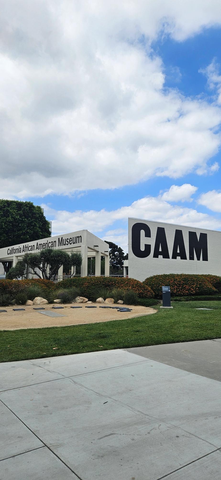 Exterior view of the California African American Museum (CAAM) building and a large "CAAM" sign, surrounded by landscaped grounds under a partly cloudy sky.