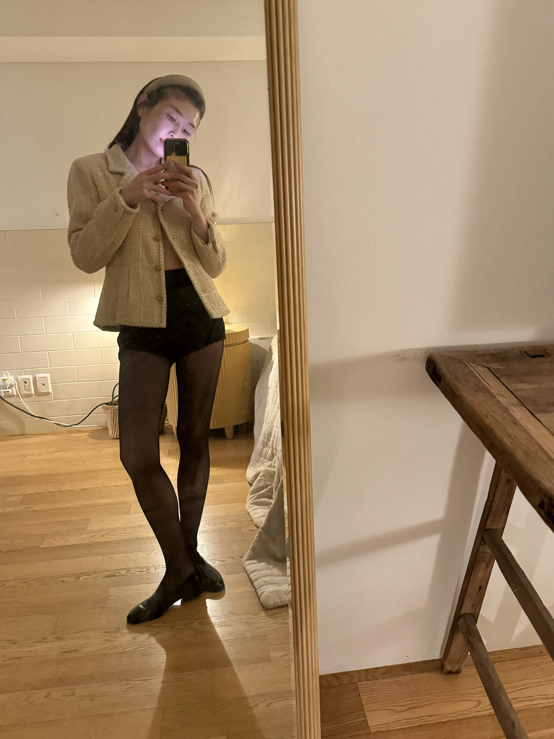 A person in a tweed jacket, black shorts, and sheer tights takes a mirror selfie.